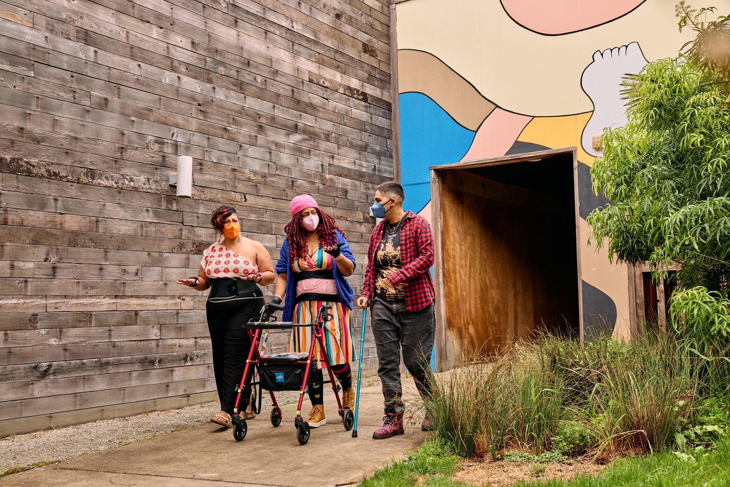 Three masked disabled people of color walk and talk. One uses a rollator, one uses a cane, and one wears wrist braces. They wear expressive, colorful outfits.