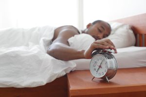“8609141689_ff923d2934_k.jpg” by Navy_NADAP is licensed under CC BY-NC-ND 2.0 Image showing a person reaching out from a bed to shut off an alarm clock on side table