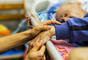 “hospice-1761276_1280.jpg” by truthseeker08 is licensed under CC0 Image showing elderly man, laying in bed, holding hands of person who is out of view