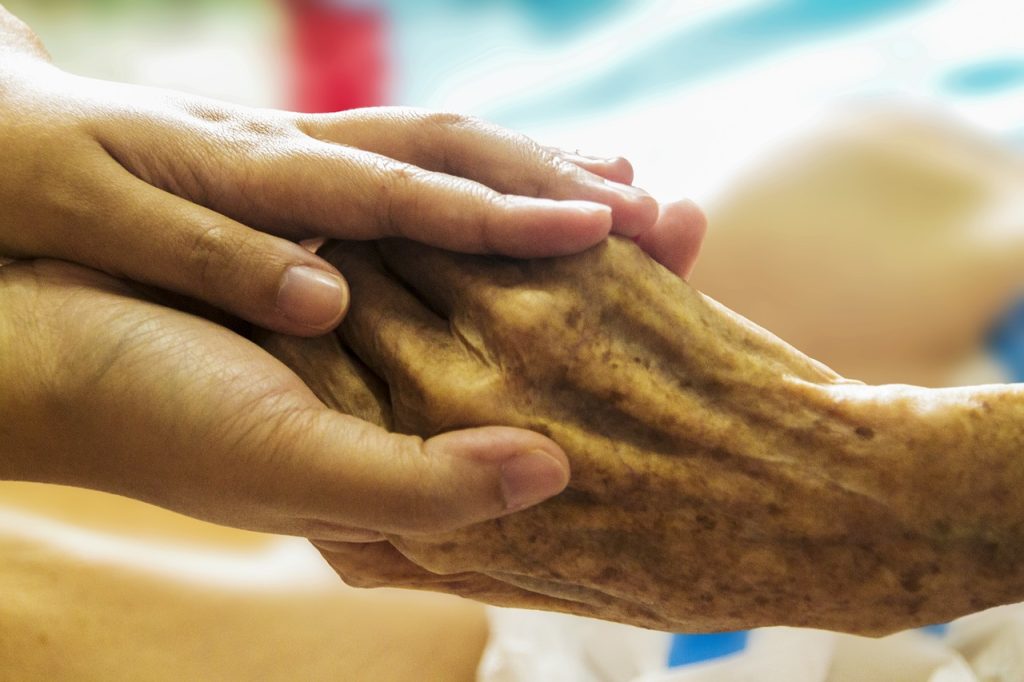 “hospice-1793998_1280.jpg” by truthseeker08 is licensed under CC0 Photo showing closeup of a younger hand holding an elderly one