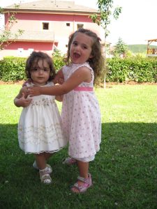 Image of two toddlers, approximate ages 2 and 3 years old, standing in front of a building in floral sundresses