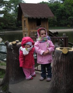 Image of two toddlers - aged approximately 2 and three years old - in pink jackets and scarfs standing in front of a pond with a small boat house in the immediate background