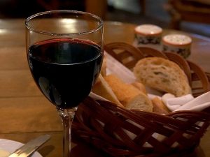 Image of a glass of red wine in front of a basket of Italian bread on a wooden table.