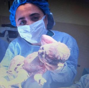 Image of a woman in surgical mask and gown holding a newly born infant