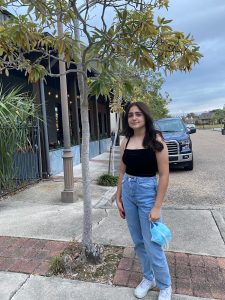 An image of an adolescent young girl with dark hair, standing on the sidewalk of a street with trees and a vehicle in the background