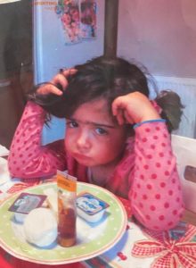 Image of a young girl in a pink polka-dotted blouse, seated at a table with a plate of food before her