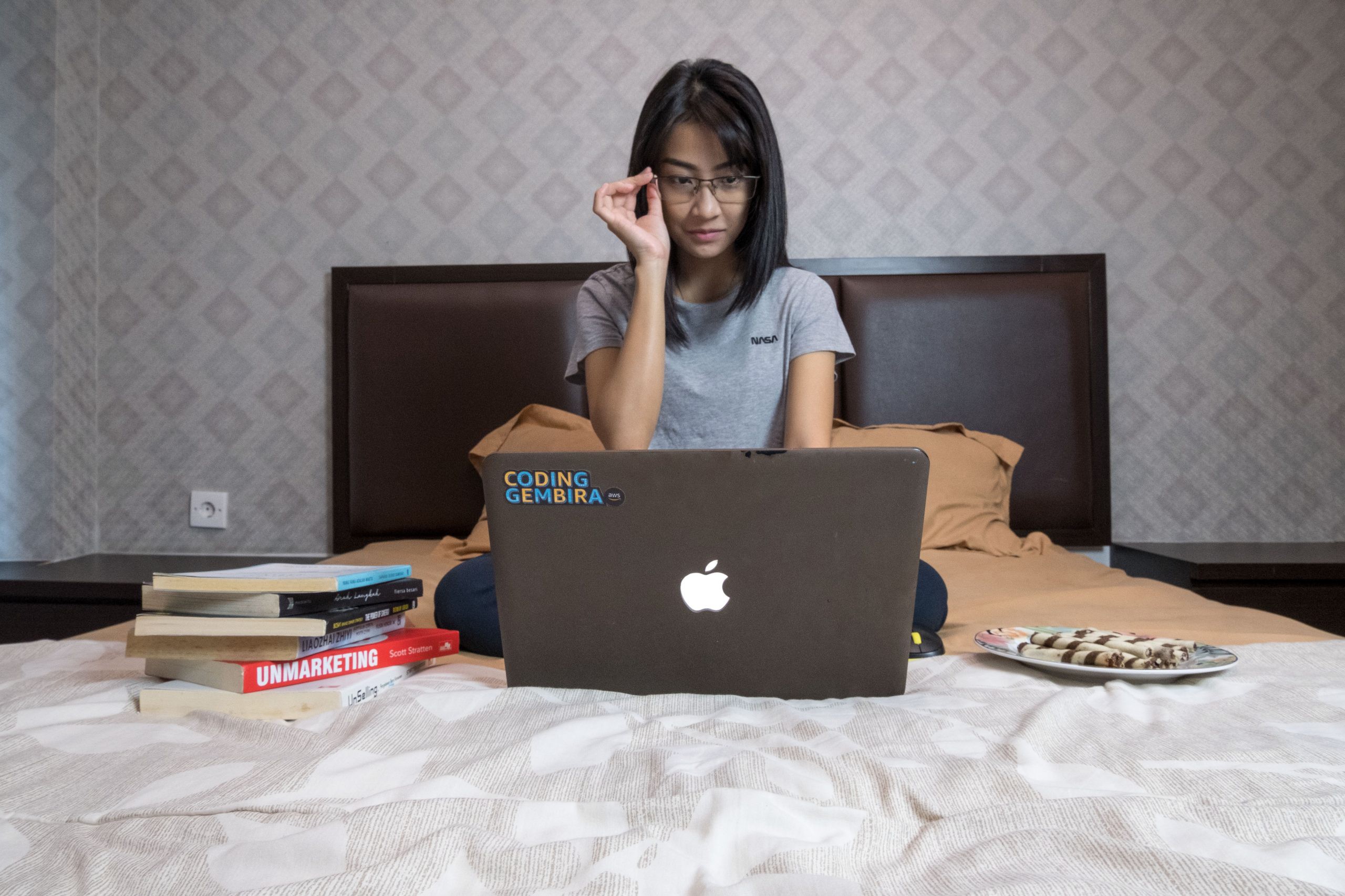 lady sitting on a bed while looking at a laptop with books on the side of her