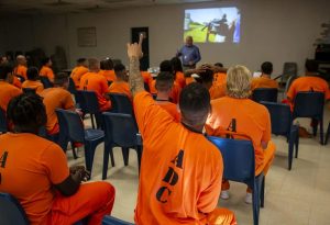 A group of inmates in orange uniforms labeled "ADC" sit in rows of blue chairs, facing a person giving a presentation with a projected image on the wall.