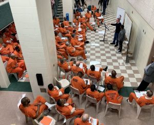 A group of inmates in orange uniforms seated in a semi-circle, listening to two presenters standing at a podium.