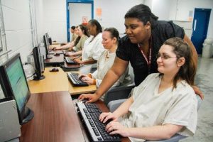 Women in white uniforms using desktop computers in a row with an instructor assisting them.
