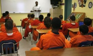 A classroom with inmates in orange uniforms seated at desks facing an instructor by a whiteboard.