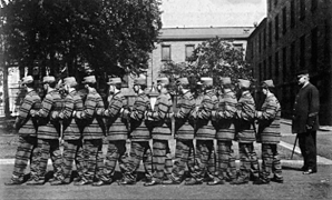 A vintage photo of men in striped uniforms standing outdoors in formation, with a guard nearby and buildings in the background.