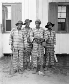 Four individuals in striped outfits holding tools, standing in front of a white building with open windows.