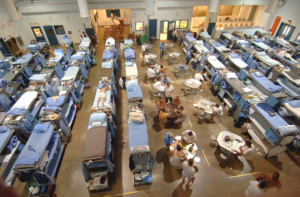Interior of a dormitory-style room with rows of bunk beds and groups of people seated at tables.
