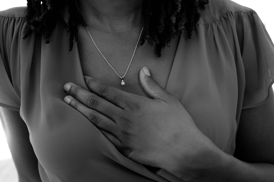 A Black woman holds her hand to her chest, wearing a necklace and blouse. The black-and-white image conveys emotion or reflection.