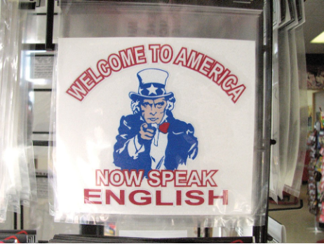 A sign on a wall that says welcome to America. Now Speak English with a man in patriotic regalia in the center.