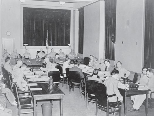 Group of people in a military commission, seated in chairs around a number of tables arranged in a U shape.