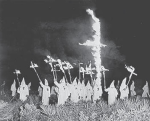 Ku Klux Klan members surrounding a large cross in the ground that is on fire while holding small burning crosses in the air.