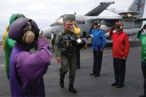 George W. Bush in a flight suit stepping out of a plane onto an aircraft carrier. Personnel stand on either side and salute him.