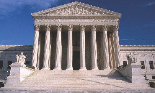 Supreme Court building. In the foreground, a set of stairs is bracketed by statues on either side, leading up to a portico. The portico has a roof supported by several tall columns.