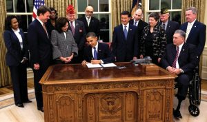 12 people standing around Barack Obama, who is seated at a desk and signing a piece of paper.