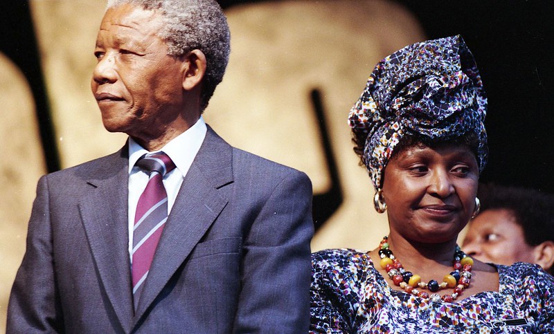 Color photograph of Nelson Mandela in suit and tie with his wife, Winnie Mandela in floral printed clothes and headwrap and multi-colored necklace