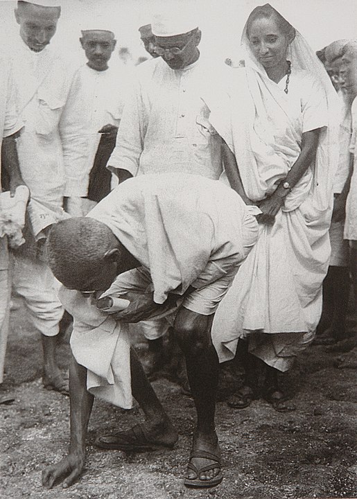 Gandhi bending over to pick up salt on the beach at the end of the Salt March with a group of people behind him