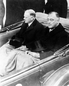 Herbert Hoover (left) and Franklin D. Roosevelt seated in a car, holding top hats, with laps covered by blanket