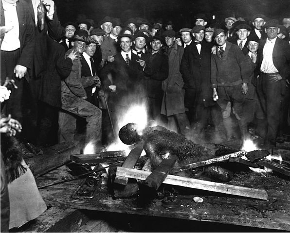 The charred remains of Will Brown on top of a burning fire while a group of men surround the corpse, many smiling for the photograph
