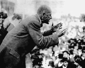Eugene Debs speaking to a crowd, leaning over a bannister with arms partly raised