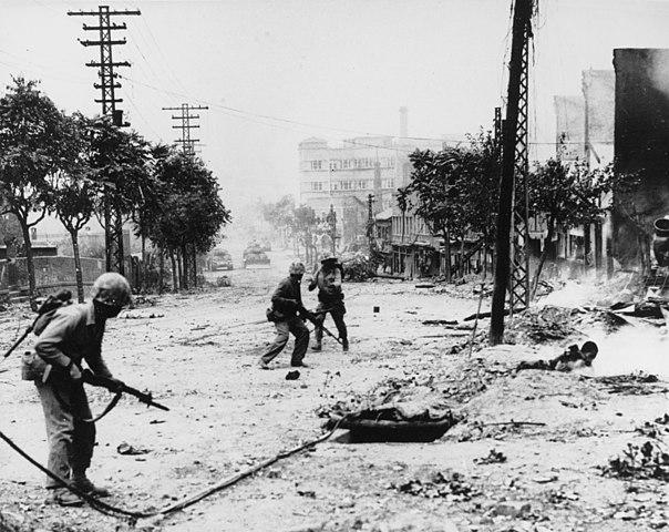 U.S. Marines engaged in street fighting during the liberation of Seoul. Note M-1 rifles and Browning Automatic Rifles carried by the Marines, dead Koreans in the street, and M-4 "Sherman" tanks in the distance