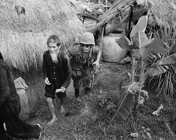 US Marine in military fatigues, helmet, and sunglasses holding a gun moves an old man with long beard suspected of being a member of the NLF from a grass covered hut