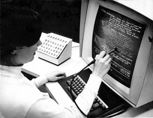 Woman sitting at computer using a Hypertext Editing System (HES) console including display unit with keyboard, light pen and function keys.