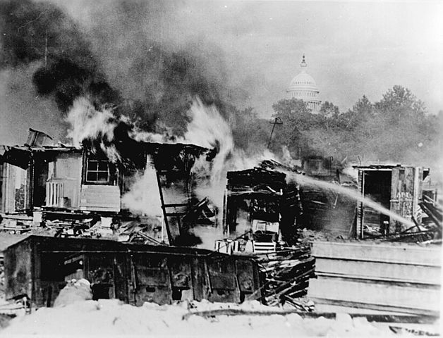 Shacks burning while stream of water being used to put out fire with view of top of Capitol building in background