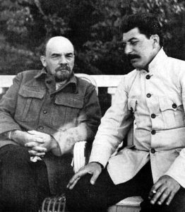 Lenin and Stalin seated next to each other on a bench, Lenin, on left, looking into camera with hands resting on lap, Stalin, on right, turned slightly towards Lenin