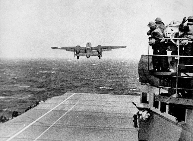 Plane takes off from the deck of the USS Hornet over open ocean while naval personnel watch