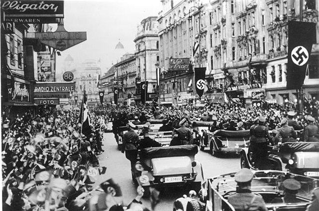 Crowds in street cheering on Nazi soldiers in vehicle parade in Vienna with large flags hanging from buildings bearing swastikas