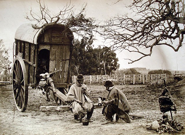 A group of Argentine gauchos, drinking mate and playing guitar, in front of a covered wooden wagon