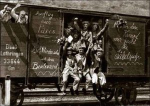 Nine German soldiers hanging out of supply train with their arms raised some with flowers others waving or raising their clenched fists. Messages on the car spell out (approximately): "Trip to Paris", "See you later on the Boulevard", "[obscured by flowers] the fight" (The obscured part most likely reads "Auf in [den Kampf]" which means "Into battle"), "my sword tip is itching".