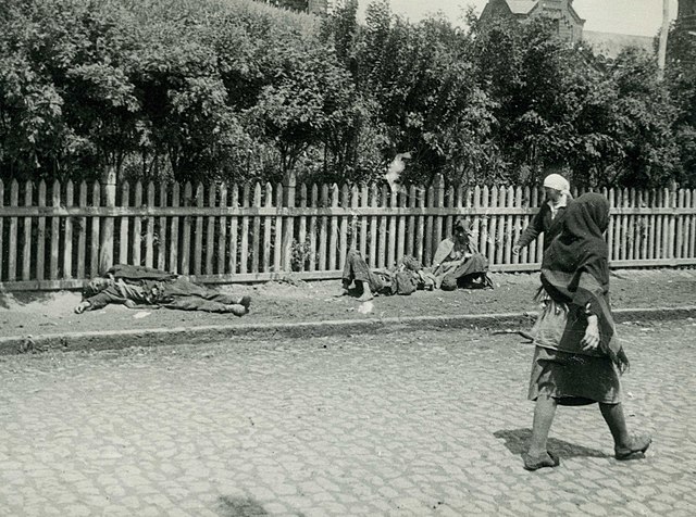 Starving peasants lying on sidewalk in front of fenced yard or garden while women on the street walk by
