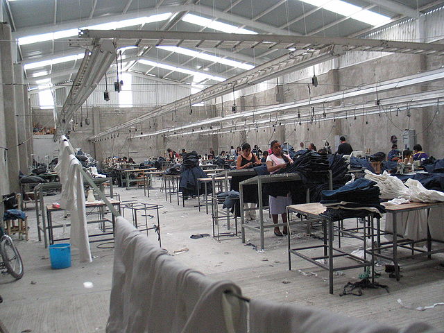 Women stand at desks making clothes in a large room