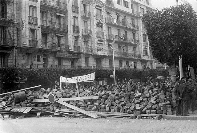 Street barricade of stone blocks, wooden boards, and other large detritus with sign that appears to read "Vive Massu" with apartment building in background