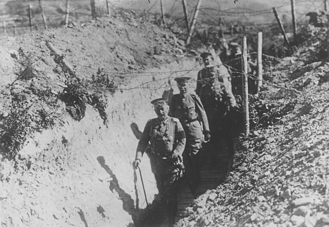 Kaiser Wilhelm walking in a trench followed by men in military dress with barbed wire fences hanging over the top of the trench