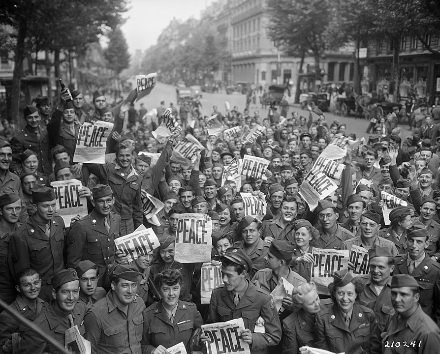 Large group of Allied military personnel in the street celebrating V-J Day holding up newspapers with the headline "Peace"