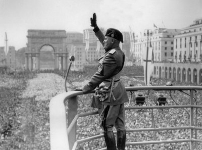 Benito Mussolini in military dress raises gloved right hand in Roman Salute to large crowds beneath him