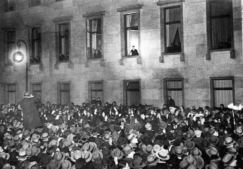 Adolf Hitler illuminated at a window of building greeting a large crowd in the street on the evening of his inauguration as chancellor