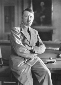 Photographic portrait of Adolf Hitler in suit leaning on desk with arms crossed looking directly into camera