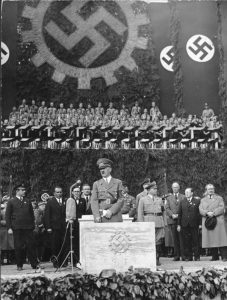 Hitler standing at white podium with swastika with group of people behind him, above are flags and a large wall mural with swastikas