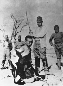 Chinese POW with torn shirt kneeling before Japanese officer with sword raised for beheading while others look on