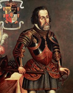 Hernán Cortés in black and leather armor holding sword hilt in right hand and long, brown cylindrical object in other